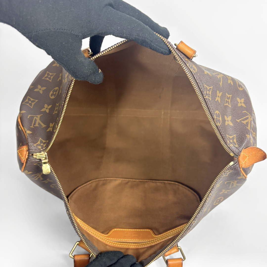 An open Louis Vuitton monogram handbag shown from the inside, with a black gloved hand holding the zipper open. The bag features the classic brown and tan monogram pattern, tan leather trim, and gold-tone hardware on a plain white background.