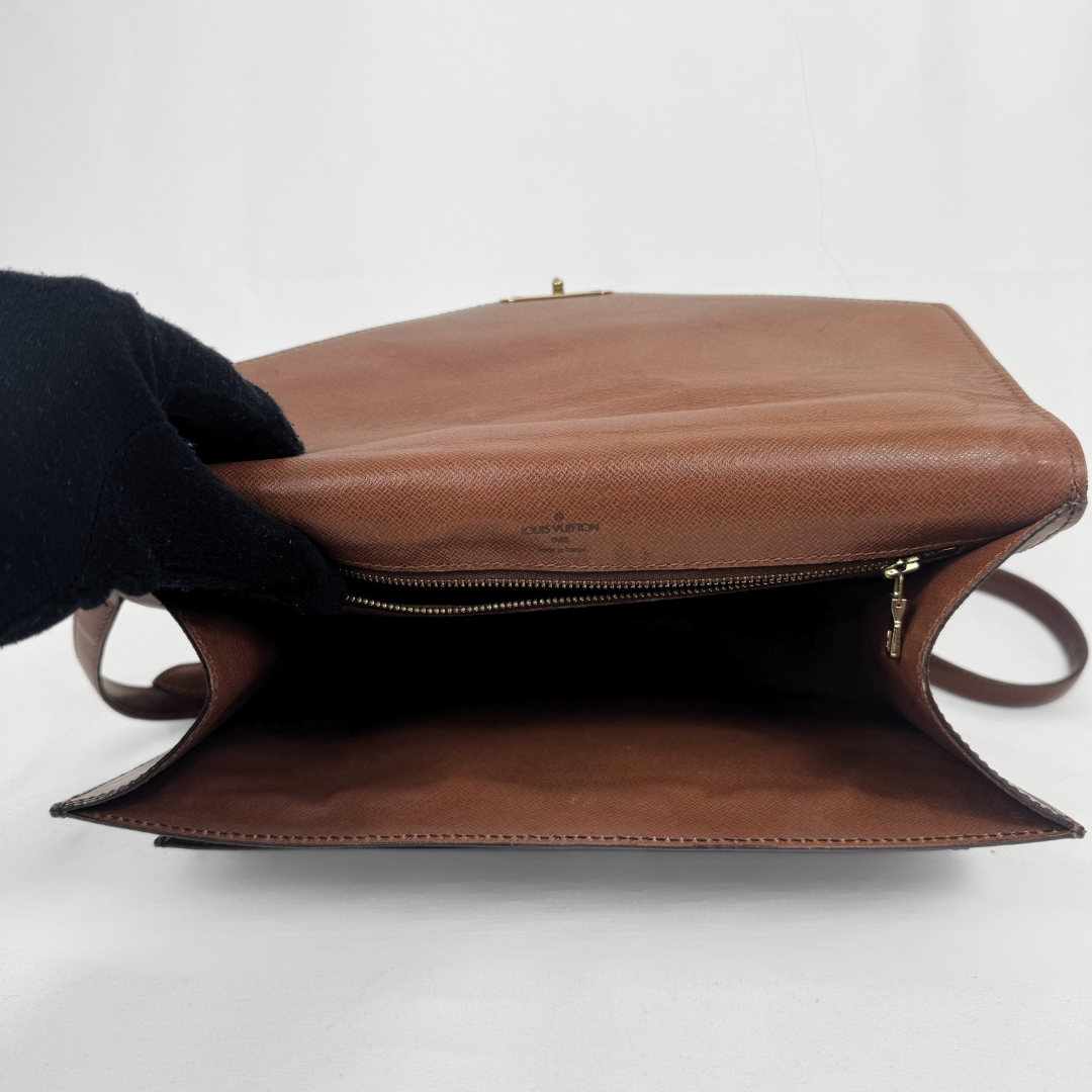 A brown leather shoulder bag shown open, with a black gloved hand holding the flap. The interior is dark, and the exterior features a tan leather body with gold-tone hardware and a small embossed logo on the front.