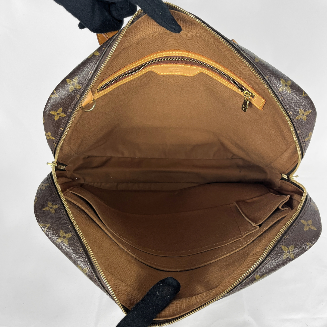An open Louis Vuitton monogram handbag shown from above, revealing a brown interior with gold-tone zippers and a small zip pocket. Gloved hands are holding the bag open against a plain white background.