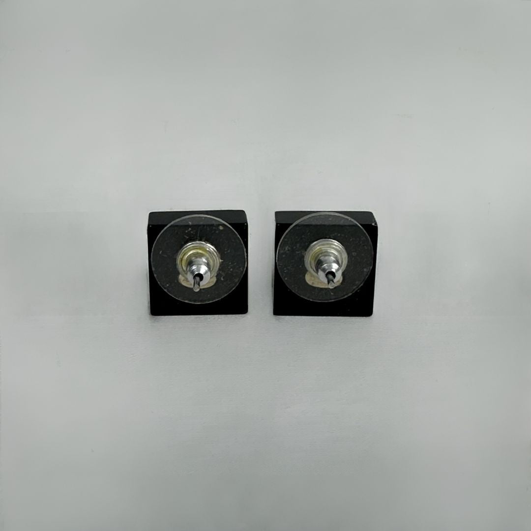 A pair of black square stud earrings with circular clear resin tops and silver-tone metal centers, shown on a plain light gray background.
