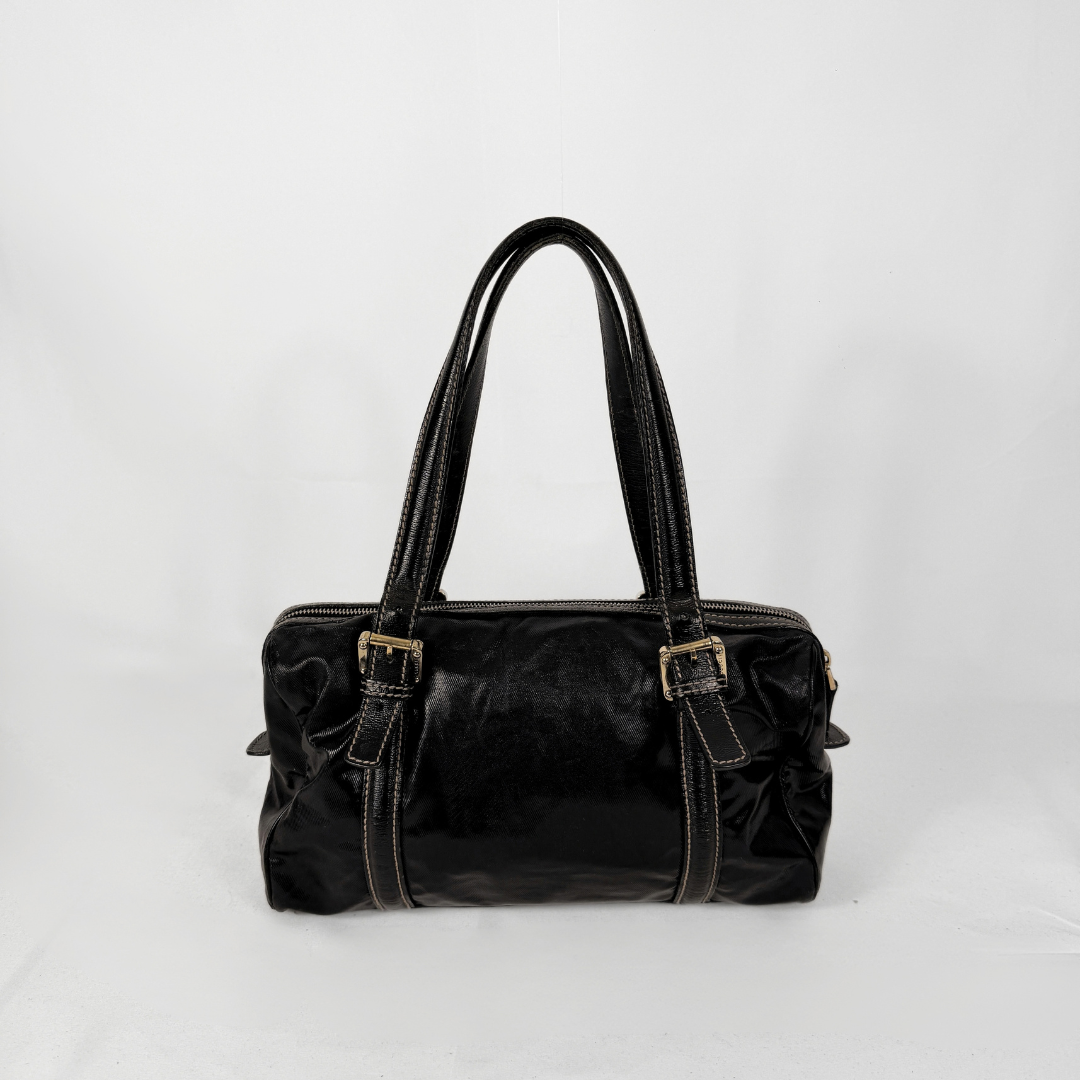A black patent leather handbag with double top handles, gold-tone buckle details, and contrast white stitching, photographed against a plain white background.