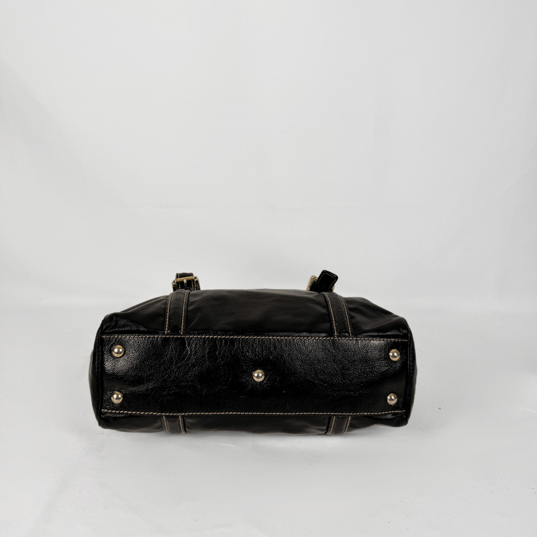 A black leather handbag shown from the bottom front view, featuring gold-tone metal studs and a black leather strap, set against a plain light gray background.