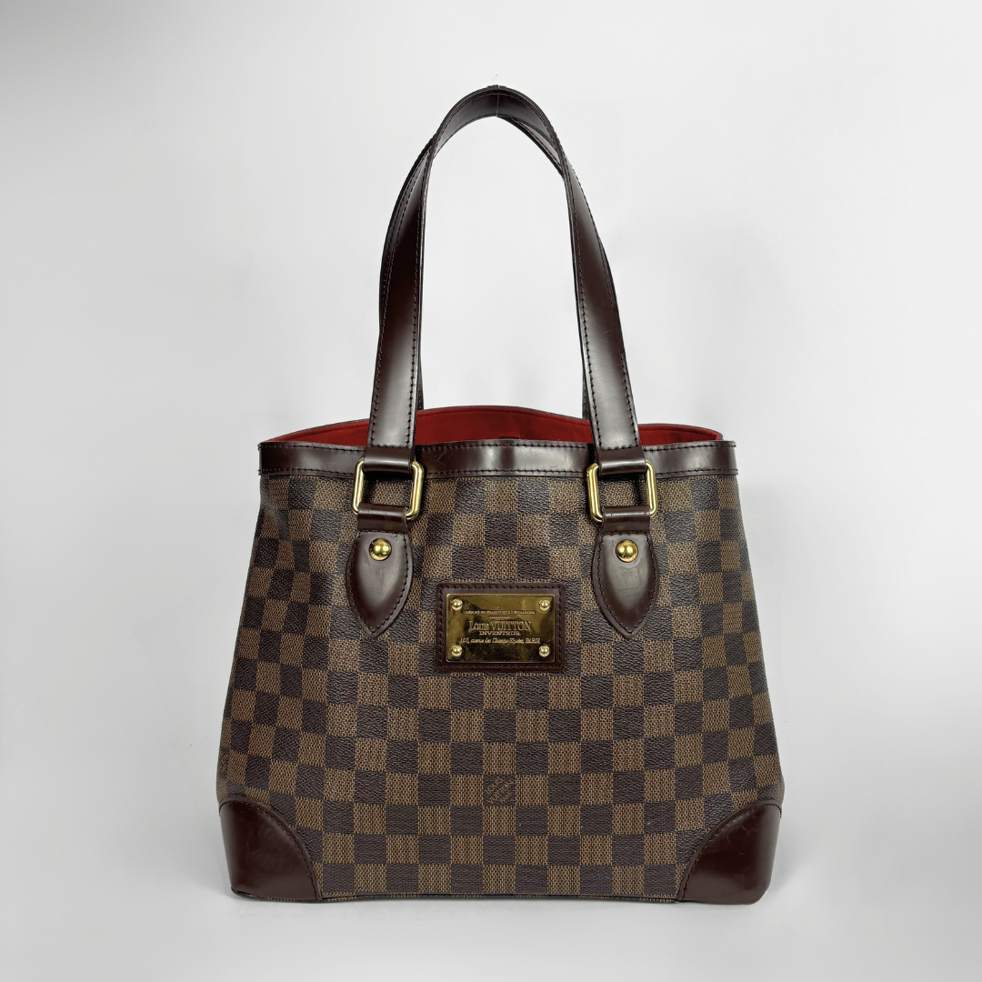 A Louis Vuitton handbag in the Damier Ebene checkered pattern, featuring dark brown leather handles and trim, gold-tone hardware, and a red interior lining, photographed against a neutral light gray background.