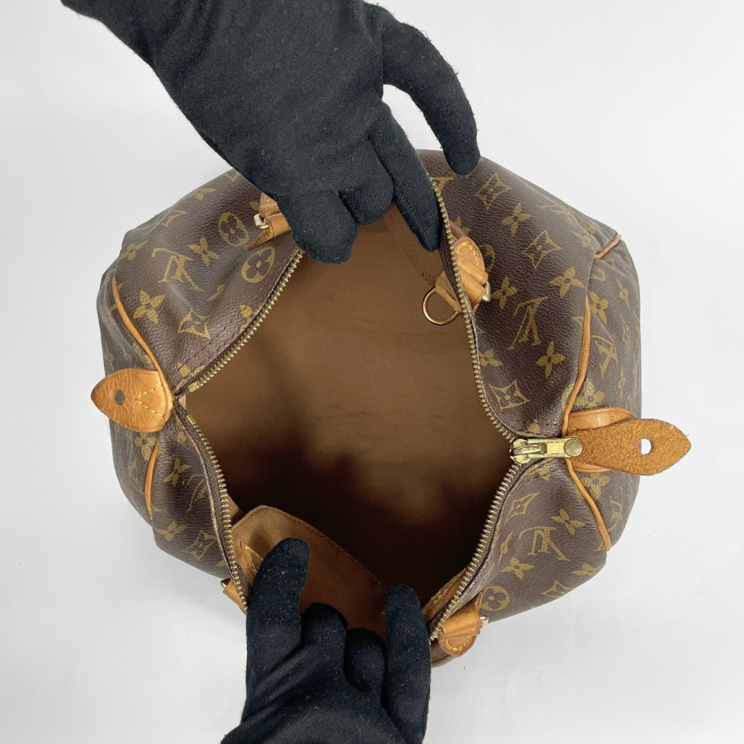 A Louis Vuitton monogram canvas handbag shown open from above, with black gloves holding the zipper area to display the interior. The bag features the classic brown and tan monogram pattern with gold-tone hardware.