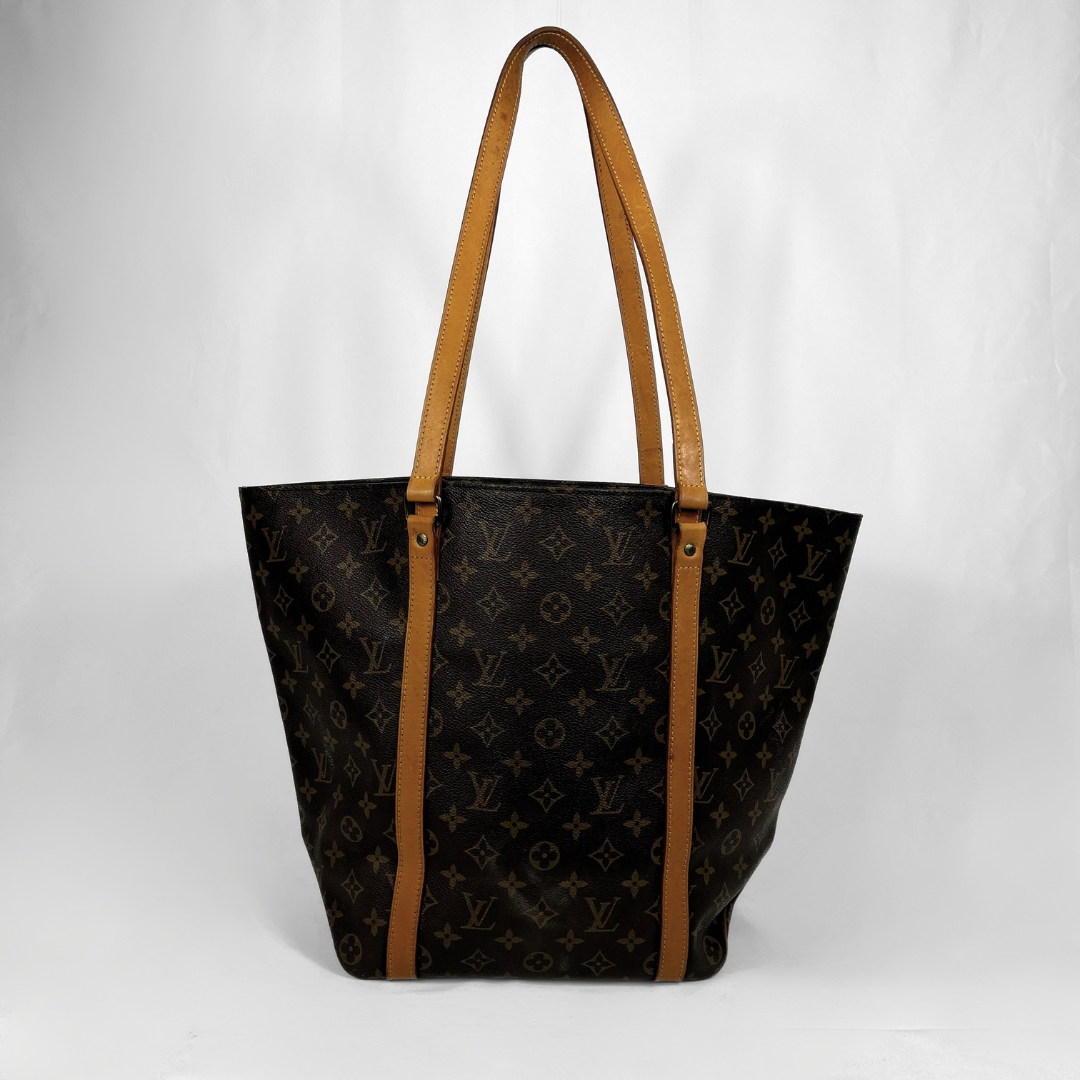 A Louis Vuitton tote bag in black monogram canvas with tan leather shoulder straps and trim, photographed against a plain light gray background.