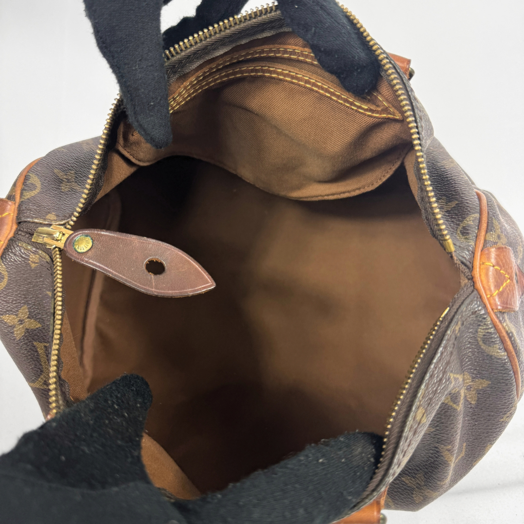 An open Louis Vuitton monogram bag shown from the inside, with black-gloved hands holding it open. The bag features the classic brown coated canvas with tan monogram motifs, gold-tone zippers, and a brown leather interior lining.