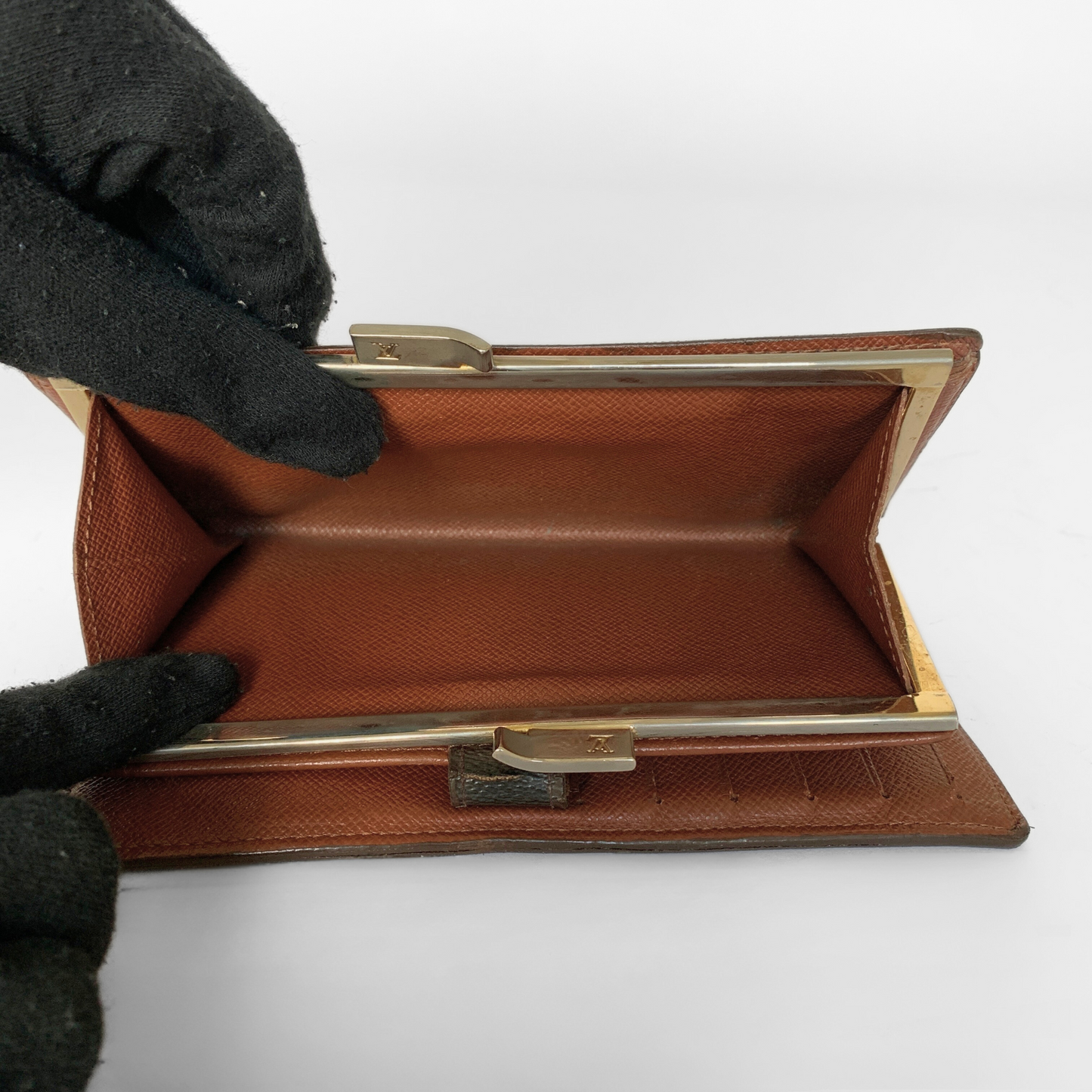 A brown textured leather clutch wallet with a gold-tone metal frame, shown open with a black-gloved hand holding it against a white background.