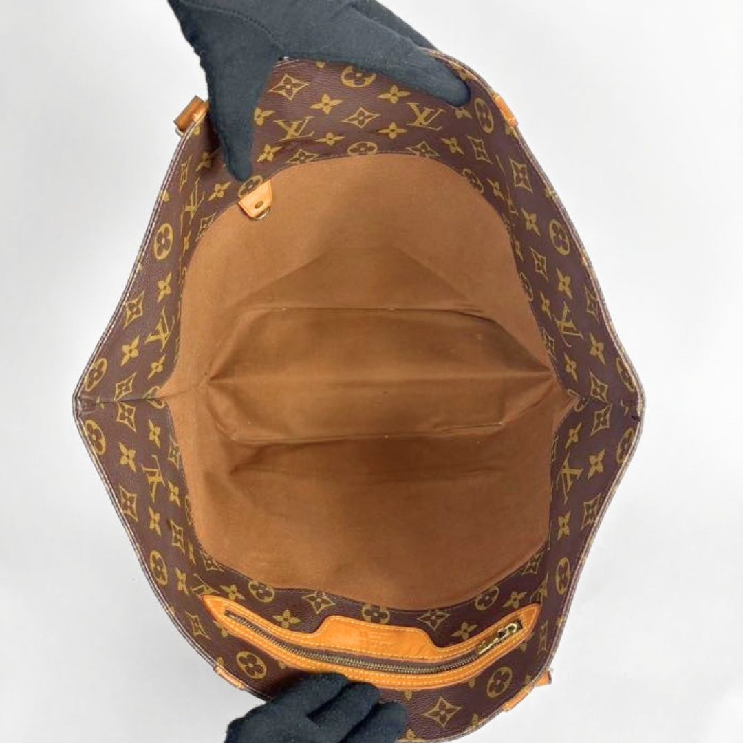 The interior of a Louis Vuitton monogram canvas tote bag, shown from above, with a brown fabric lining and a zippered compartment. Gloved hands are holding the bag open against a plain white background.