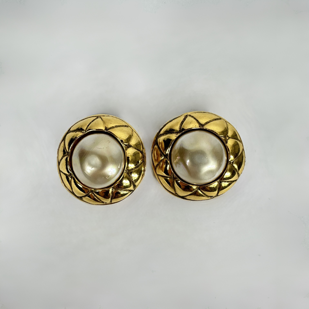 A pair of gold-tone round stud earrings with textured, diamond-patterned outer rings and smooth, lustrous white faux-pearl centers, displayed on a light gray background.