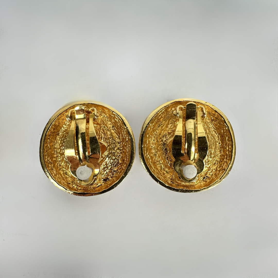 A pair of gold-tone round clip-on earrings shown from the back, displaying the textured enamel surface and metal clasp mechanism, set against a plain light gray background.