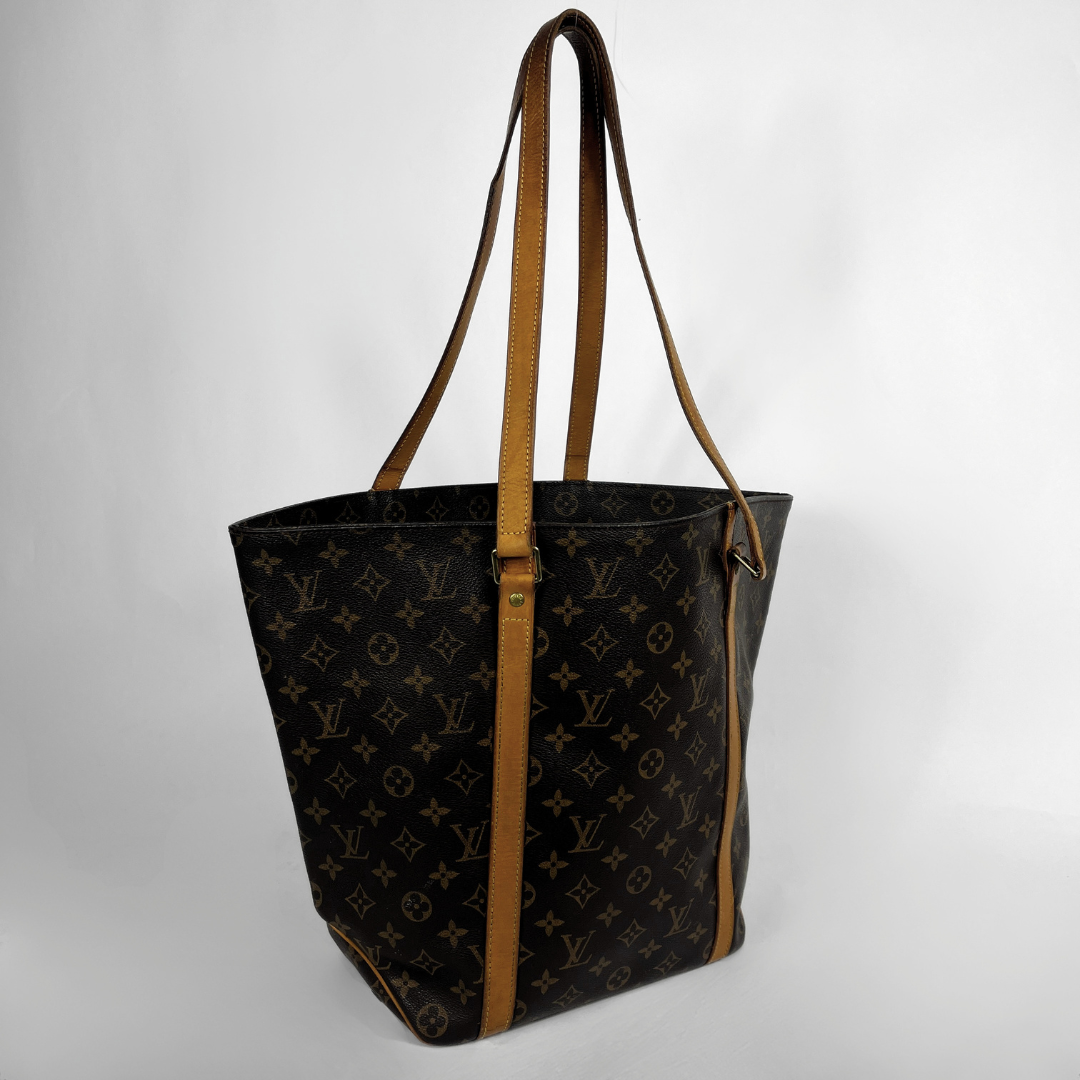 A Louis Vuitton tote bag in dark brown monogram canvas with tan leather shoulder straps and trim, photographed against a light gray studio background.