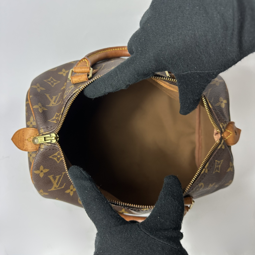 A Louis Vuitton monogram canvas handbag shown open from above, with black-gloved hands holding the zipper area to display the interior. The bag features the classic brown and tan monogram pattern with tan leather trim and gold-tone hardware.