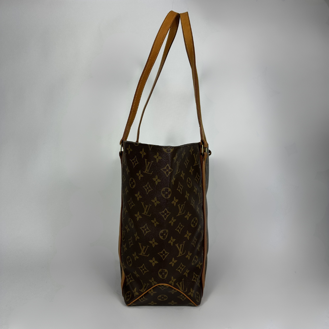 A Louis Vuitton monogram tote bag in brown coated canvas with tan leather shoulder straps, photographed against a neutral gray studio background.