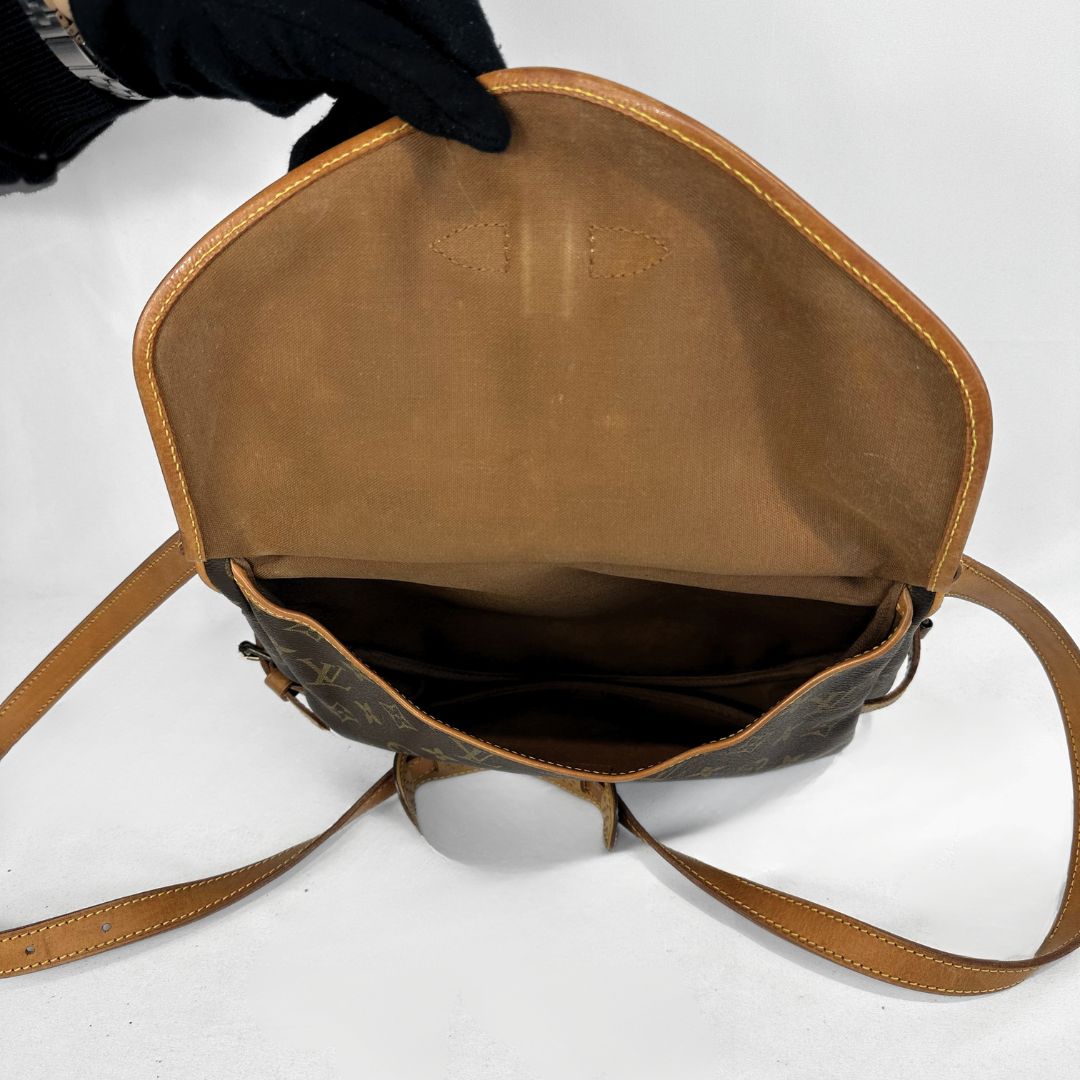 An open Louis Vuitton shoulder bag in brown monogram canvas, showing the tan leather interior lining and strap, held open by a hand wearing a black glove against a white background.