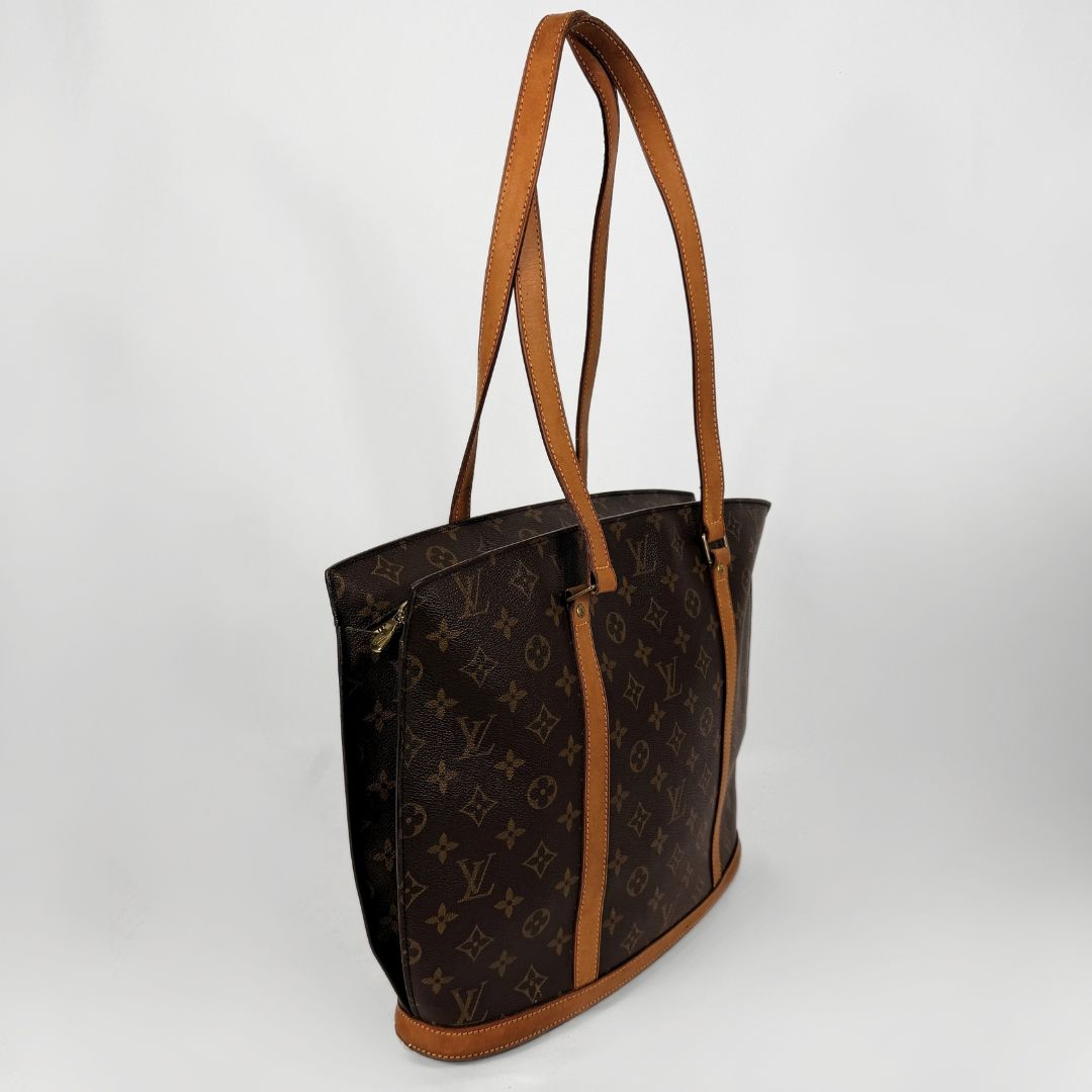 Louis Vuitton monogram canvas tote bag with brown leather trim and shoulder straps, shown on a light gray background.