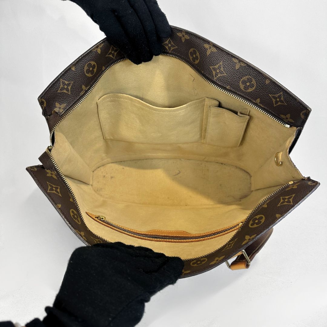 Louis Vuitton monogram canvas shoulder bag shown open, with black gloved hands holding the edges to display the interior. The bag features the classic brown and beige monogram pattern, a beige interior lining, and gold-tone hardware.