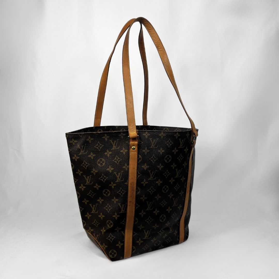 A Louis Vuitton monogram canvas tote bag with brown leather shoulder straps and trim, photographed against a white studio backdrop.