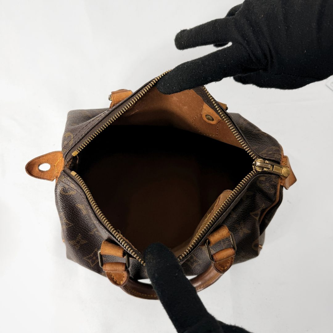 A Louis Vuitton monogram canvas handbag shown open from above, with a black gloved hand pointing toward the interior. The bag has dark brown coated canvas with tan leather trim and gold-tone hardware on a plain white background.