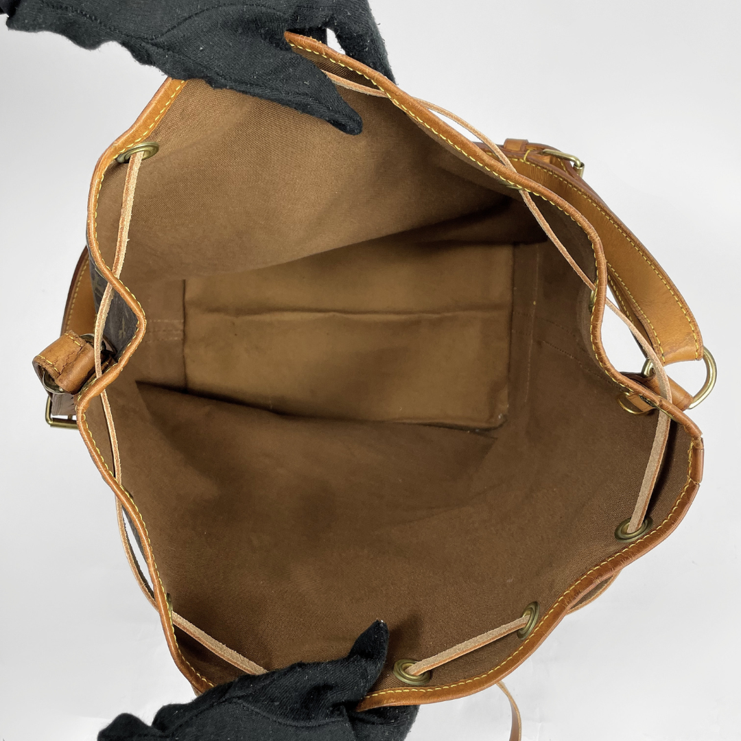 An open tan leather and canvas bucket bag shown from the inside, with a brown suede-like lining, drawstring closure, and black-gloved hands holding it against a light gray background.