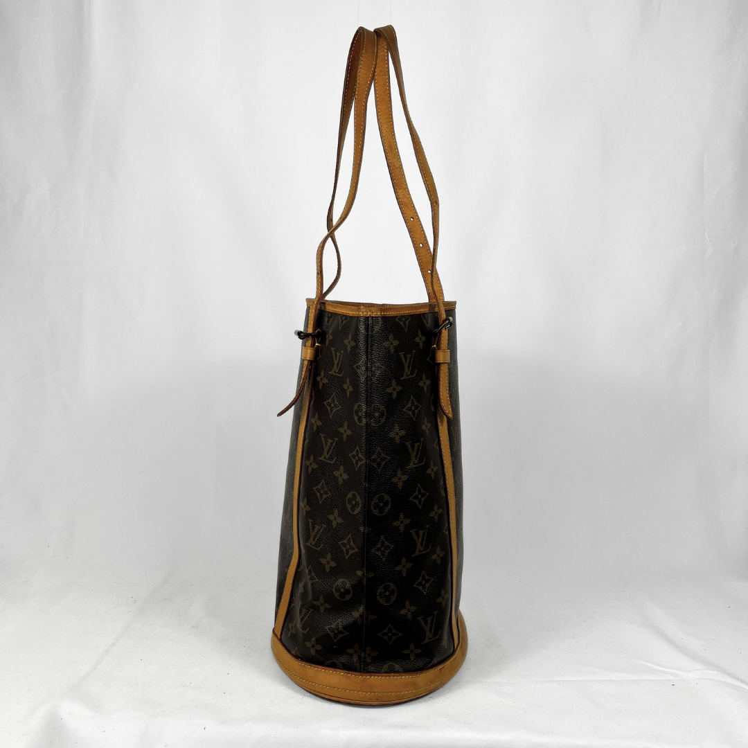 A Louis Vuitton monogram canvas tote bag with tan leather trim and shoulder straps, standing upright against a white fabric backdrop.