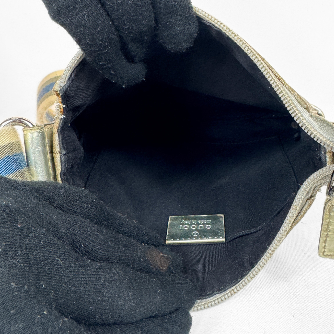 An open black handbag with a black interior lining and a silver-tone zipper, shown with black gloves holding it open. A small metal brand plaque is visible inside, and part of a patterned strap and gold-tone hardware appear at the edges.