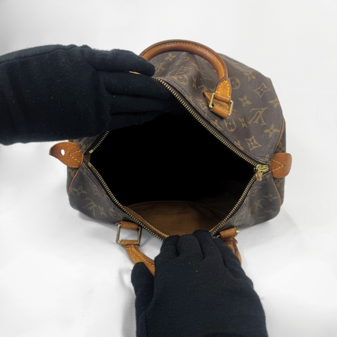 A Louis Vuitton monogram Speedy bag shown open, with black gloved hands holding the handles to display the interior. The iconic brown monogram canvas and tan leather details are clearly visible against a plain white background.