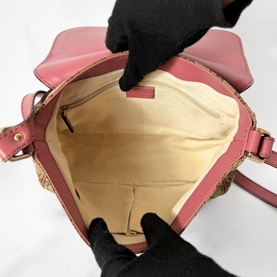An open pink crossbody bag with a beige interior lining, held open by two hands wearing black gloves against a white background.